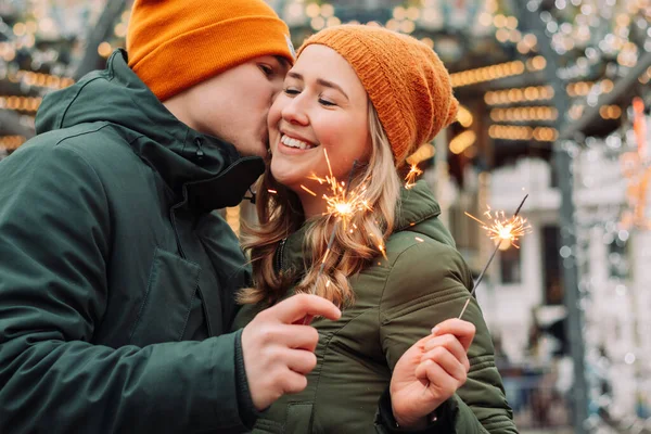 Young attractive couple hug and kiss outdoors with the sparklers in their hands celebrating New Year and Christmas. Winter holidays festival mood. Man and woman feel happy. Love and happiness concept