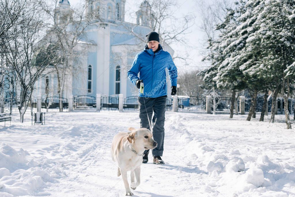 Dog's owner with his pet having fun in the park on a snowy winter's day. Man and a labrador retriever run and being active outdoors among piles of snow. Friendship and togetherness concept