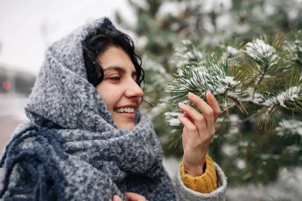 Smiling young woman stands in a frosty snowy winter park. Female wearing a grey coat and blue scarf having fun among fir trees covered with snow. Winter holidays, Christmas and festive mood concept