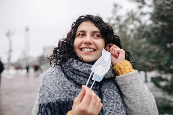 Young woman takes on the medical sterile mask at a winter snowy park on a cold frosty day. Coronavirus spread prevention druing pandemic outbreak. Personal protective equipment and health concept