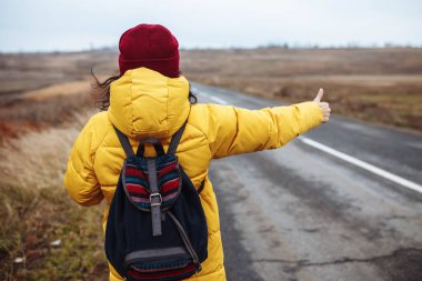 Sırt çantalı, sarı ceketli ve kırmızı şapkalı bir kadın turist yolda bir araba yakalıyor. Genç bir kadın kışın veya sonbahar sonlarında seyahat eder. Otostop, gezi, baş parmak yukarı, seyahat konsepti.