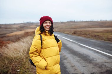 Sırt çantalı, sarı ceketli ve kırmızı şapkalı bir kadın turist portresi yolda duruyor. Genç bir kadın kışın veya sonbahar sonlarında seyahat eder. Otostop, gezi ve seyahat konsepti