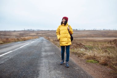 Sırt çantalı, sarı ceketli ve kırmızı şapkalı bir kadın turist yolda yürüyor. Genç bir kadın kışın veya sonbahar sonlarında seyahat eder. Otostop, gezi ve seyahat konsepti