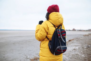 Sırt çantalı genç bir turistin engin kış vadisinin ovalarında yan yana yürürken görüntüsü. Sarı ceketli ve kırmızı şapkalı bir bayan yolcu. Otostop, seyahat konsepti