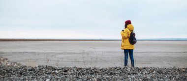 Sırt çantalı genç bir turistin engin kış vadisinin ovalarında yan yana yürürken geniş açılı görüntüsü. Sarı ceketli ve kırmızı şapkalı bir kadın. Otostop, seyahat konsepti