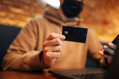 Cep telefonu, dizüstü bilgisayar ve kredi kartıyla alışveriş ve online ödeme. Covid-19 sırasında siyah tıbbi maske takan adam bir kafede oturur ve internetten mal alır. Yeni normal ve teknoloji kavramı