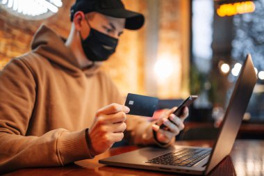 Cep telefonu, dizüstü bilgisayar ve kredi kartıyla alışveriş ve online ödeme. Covid-19 sırasında siyah tıbbi maske takan adam bir kafede oturur ve internetten mal alır. Yeni normal ve teknoloji kavramı