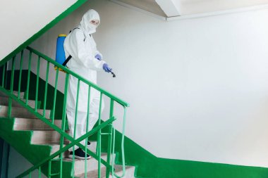 Koruyucu giysi giyen hijyenik profesyonel işçi apartman girişini dezenfekte ediyor. Yerleşim bölgelerinde koronavirüs önleme önlemleri. Yeni normal ve Covid-19 yayılma konseptini durdur