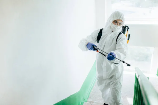 Koruyucu giysi giyen hijyenik profesyonel işçi apartman girişini dezenfekte ediyor. Yerleşim bölgelerinde koronavirüs önleme önlemleri. Yeni normal ve Covid-19 yayılma konseptini durdur