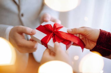 Sevgililer Günü konsepti. Genç, romantik bir çift ellerinde bir hediye tutuyorlar. Önünde de çelenk ışıkları var. Kırmızı kurdeleli beyaz hediye kutusu. 14 Şubat kutlamaları.