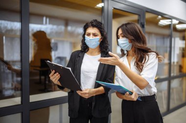 İki iş kadını, steril maske takarak iş meselelerini ofiste tartışıyor. Koronavirüs salgını karantinasında çalışıyor. Yeni normal, sağlık ve güvenlik önlemleri kavramı