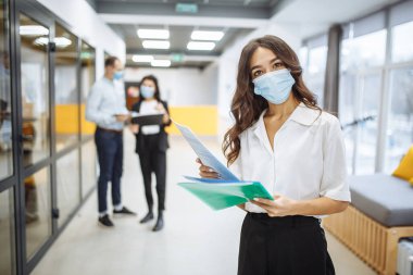 Sağlık maskesi takan genç bir iş kadınının portresi. Ofis koridorunda evrakları, iş eğilimlerini ve beklentileri kontrol ediyor. Yeni normal, sağlık ve koronavuris yayılma önleme kavramı