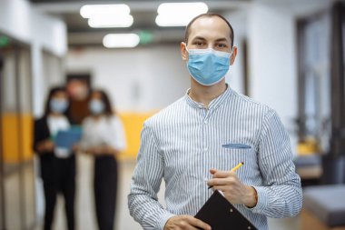 Tıbbi maske takan zarif bir işadamı elinde tabletle ofis koridorunda duruyor. Coronavirus önleme önlemleri, yeni normal kavram. Profesyonel bir işçinin portresi