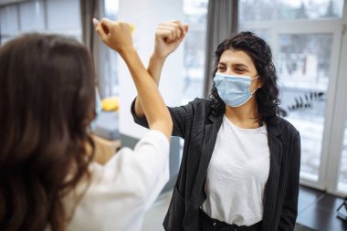 Koronavirüs salgını sırasında maske takan iki iş kadınını güvenli bir şekilde karşılamak. Dişiler bir ofis koridorunda birbirlerini selamlamak için dirseklerini çaprazlarlar. Covid-19 yayılmayı önleme, yeni normal kavram