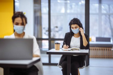 Ofis masalarında çalışan, tıbbi maske takan ve salgın sırasında güvende olan iki kadının portresi. Sağlık ve kişisel hijyen kavramı