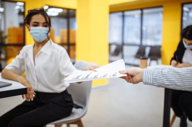 Genç meslektaşlar, Coronavuris salgını sırasında ofiste çalışan belgeleri birbirlerine veriyorlar. Tıbbi maskeler takan profesyonel işçiler iş dünyasındaki haberleri ve eğilimleri kontrol ediyorlar.