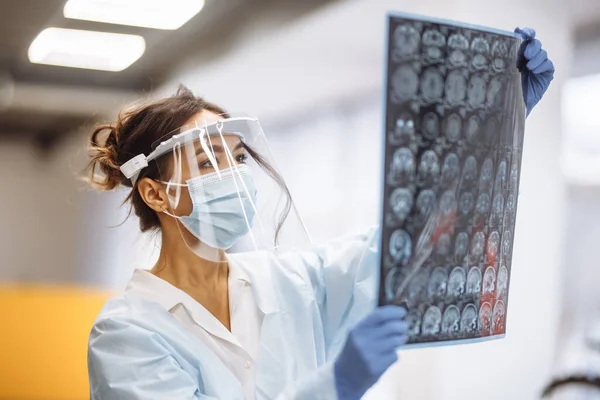 Woman doctor is checking brain MRI x-ray image of the patient at the hospital. Female medical worker wearing protective mask and white gown at the corridor of a hospital. Heath and medicine concept
