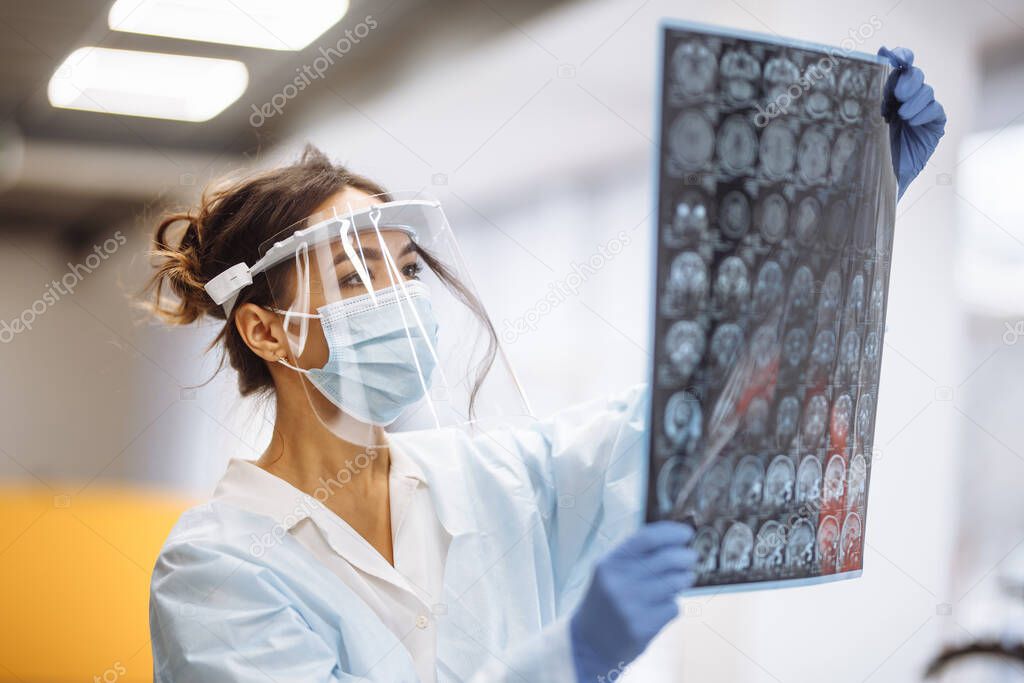 Woman doctor is checking brain MRI x-ray image of the patient at the hospital. Female medical worker wearing protective mask and white gown at the corridor of a hospital. Heath and medicine concept