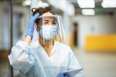 Genç hemşire Coronavirus salgını sırasında hastane koridorunda yüz kalkanını ayarlıyor. İş yerinde profesyonel tıp çalışanı portresi. Sağlık, tıp ve güvenli konsept