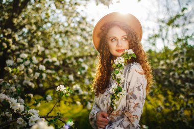 Bahar mevsiminin tadını çıkaran ve parktaki çiçek açan ağaçların arasında duran genç bir kadının portresi. Bej şapkalı ve beyaz elbiseli bir kadın mutlu çiçek ve çiçek açan ağaçlarla çevrili