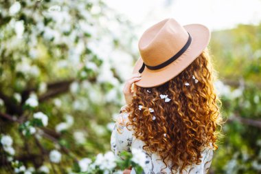 Kıvırcık saçlı, bej şapkalı, beyaz elbiseli, bahar parkındaki çiçek açan ağacın yanında duran genç bir kadının arka planı.