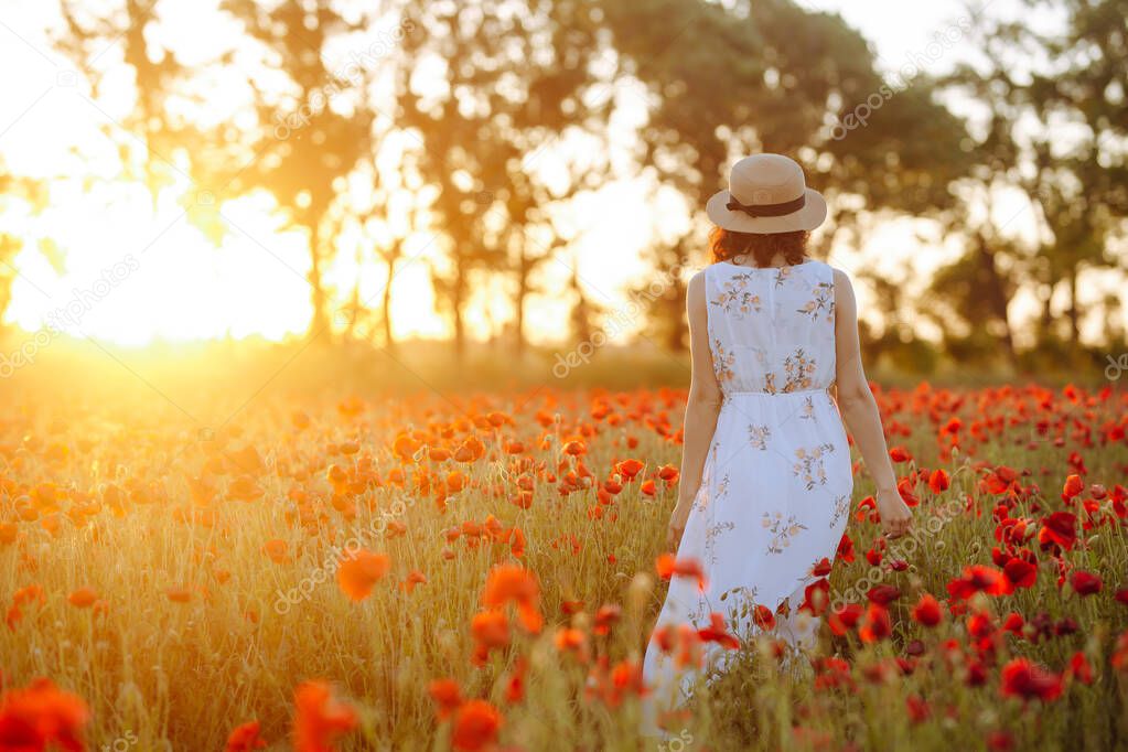 Girl wearing white dress and straw hat walks among red poppy flowers on the sunset. Young woman enjoys good waether and having fun on the field full of red flowers. Spring and nature concept
