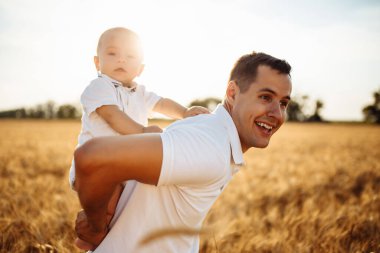 Buğday tarlasında mutlu bir baba ve oğul. Bir adam kucağında küçük bir çocuk tutuyor. Bebeğin babasının kollarında olduğu bir yaz fotoğrafı.