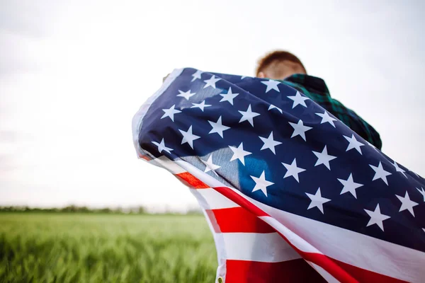 4 Temmuz. Bağımsızlık Günü. Elinde Amerikan bayrağı olan bir adam yeşil buğday tarlasının arka planında. Özgürlük kavramı. Ekose gömlekli, bayrak taşıyan genç adam.