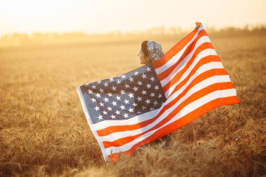 Kadın çiftçi, Amerikan bayrağıyla olgun bir buğday tarlasında güzel bir günbatımının arka planında duruyor. Rüzgarda dalgalanan bayrak. Yaz manzarası