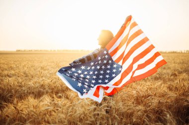 Dişi çiftçi, gün batımının arka planına Amerikan bayrağı sallıyor. Yazın buğday tarlasında bayrağı olan bir kadın. Bağımsızlık Günü. 4 Temmuz