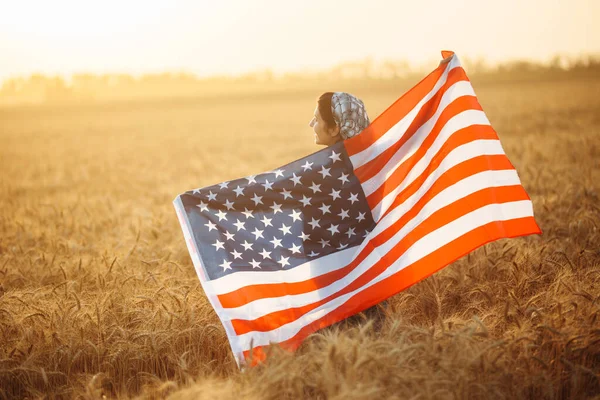 女性農家は美しい夕日を背景に熟した小麦畑にアメリカの国旗が立っています 旗は風になびく 夏の風景 — ストック写真
