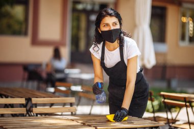 Koruyucu maskeli garson portresi ve sarı bezle ahşap masayı silen eldivenler. Tıbbi maskeli genç bir kız bir kafede mobilyaları dezenfekte ediyor. Yeni normal kavram