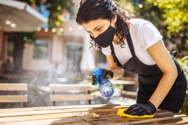 Garsonun koruyucu maskeli, eldivenli, ahşap masayı sarı bir bezle silen portresi. Tıbbi maskeli genç bir kız bir kafede mobilyaları dezenfekte ediyor. Yeni normal kavram