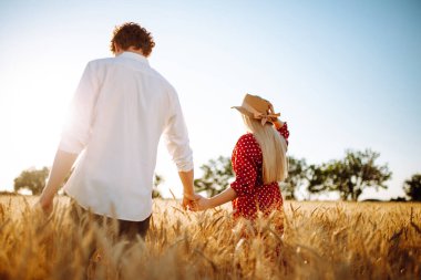 Genç bir çift gün batımında buğday tarlasında yürür. Bezelye elbiseli ve hasır şapkalı bir kız ve mavi gökyüzü arka planında beyaz gömlekli genç bir adam. Yaz manzarası. Buğday tarlasında iki aşık.