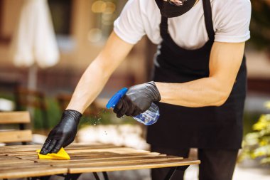 Siyah koruyucu maskeli bir garson açılmadan önce masaları dezenfektanla siler. Toz temizleme ajansı. Restoranda bir bezle masayı siliyor.