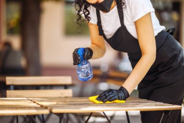 Bir garsonun eldiven ve koruyucu maskeyle bir kafe ya da restoran açılmadan önce mobilyaları dezenfekte ederken yakın plan fotoğrafı.
