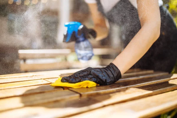 Kafe ya da restoran açılmadan önce koruyucu maske takmış bir garsonun mobilyaları dezenfekte edişinin yakın çekimi. Bir kadın işçi dezenfektan sıkar.