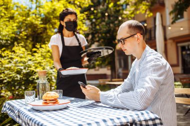 Güzel bir garson genç adama siparişini getirdi. Hamburger ve taco. Kafenin çalışanları maske ve eldiven takıyor. Yeni kurallar. Sokak terası