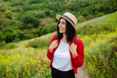 Hasır şapkalı güzel bir kız çayırda çiçeklerle birlikte ormanın arka planında yürüyor. Kırmızı kazaklı mutlu genç kadın yalnız seyahat ediyor.