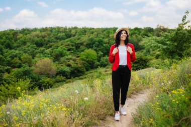 Hasır şapkalı güzel bir kız çiçeklerle çayırda yürüyor ormanın arka planında ve mavi gökyüzünde. Kırmızı kazaklı mutlu genç kadın yalnız seyahat ediyor.