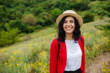 Kırmızı kazaklı ve hasır şapkalı güzel bir genç kadının portresi. Tarlada sarı çiçekli güzel bir kızın orman arka planına yakın plan fotoğrafı. Seyahat kavramı