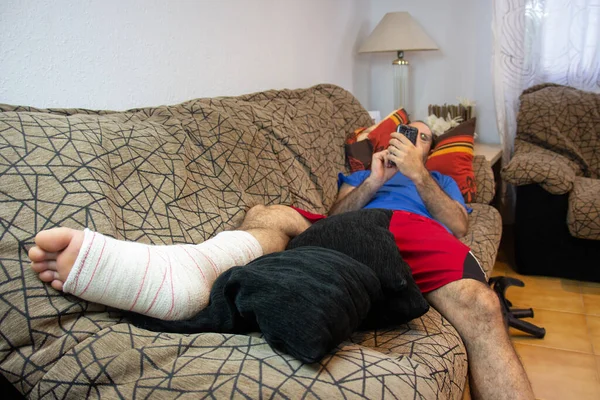 Man With Fractured Leg Sitting On Sofa Talking On Cellphone. Selective focus. Medical services. Concept of rehabilitation of people after serious physical accident injury