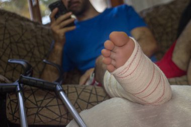 Yetişkin bir adamın bacağı alçıda ve koltuk değnekleriyle yan koltukta telefonla konuşan bir adamın yatay görüntüsü. Kazadan sonra insanların rehabilitasyonu