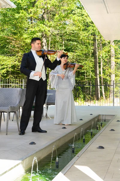 Duo of violinists. A man in a tailcoat and a woman in an evening dress play the violin near the fountain.