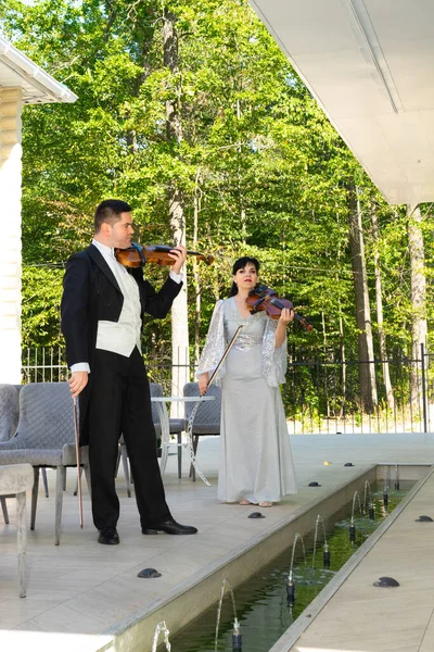 Duo of violinists. A man in a tailcoat and a woman in an evening dress are preparing to play the violin near the fountain.