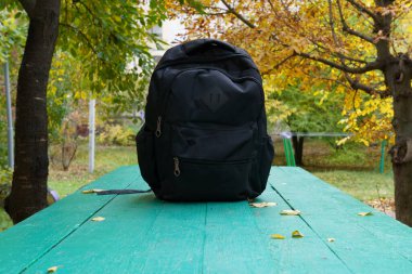 Siyah bir sırt çantası, parktaki sonbahar yapraklarıyla çevrili canlı yeşil bir masada tek başına oturur. Arkaplanda sarı yapraklı ağaçlar yer alıyor..