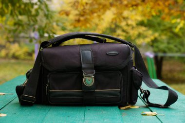 Siyah bir kurye çantası yeşil bir piknik masasında oturuyor, rengarenk sonbahar yapraklarıyla çevrili, gösterişli mevsimsel güzellikte..