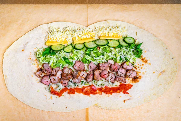 Düz bir tortilla üzerinde ızgara et, taze sebze, peynir ve sos katmanları, canlı renkler ve dokular sergilenerek bir paket hazırlanıyor..