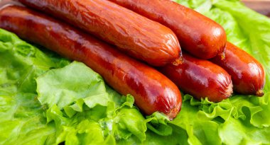 Mutfak ortamında yeşil marul yapraklarından oluşan bir yatakta dört ızgara sosis dinleniyor. Sahne yemek hazırlıklarını gösteriyor..