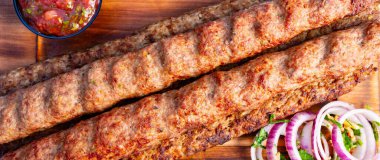 Uzun kebaplar dilimlenmiş soğanlar ve yakınlarda bir kase sosla ahşap bir tabağa konur. Bu sahne gün boyunca hareketli bir pazar ortamından..